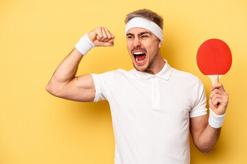 Young caucasian man holding ping pong rackets isolated on yellow background raising fist after a...