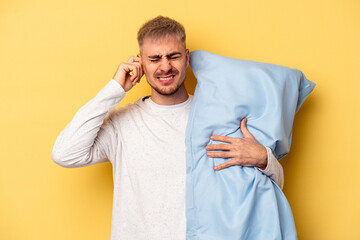 Young caucasian man wearing a pajama and holding pillow isolated on yellow background covering ears...