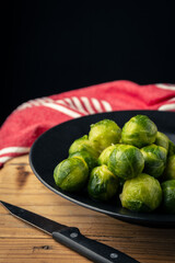 Top view of frozen brussels sprouts on black plate on wooden table with knife and red cloth, selective focus, black background, vertical