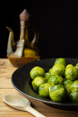 Top view of brussels sprouts on black plate on rustic wooden table with wooden spoon and oil can, selective focus, black background, vertical