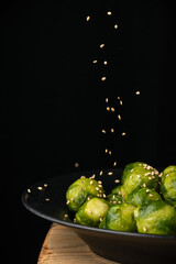 Close-up sesame falling on a plate of brussels sprouts on wooden table, selective focus, black background, vertical