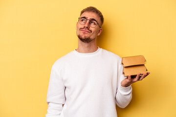 Young caucasian man holding a burger isolated on yellow background dreaming of achieving goals and...