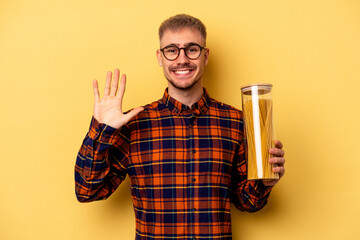 Young caucasian man holding spaghettis jar isolated on yellow background smiling cheerful showing number five with fingers.