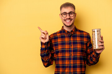 Young caucasian man holding chickpeas isolated on yellow background smiling and pointing aside, showing something at blank space.
