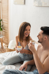 sexy woman and shirtless man smiling at each other while drinking coffee in bedroom.