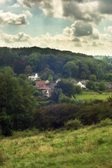 Fototapeta premium Hilly countryside with woods and houses in a valley under a blue cloudy sky in summer.