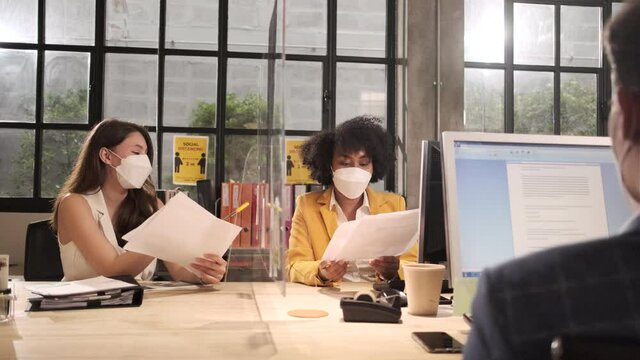 Two female coworkers team with face mask working in new normal office. COVID-19 protection by cleared partition, business workplace office, social distancing for pandemic health, disease prevention.