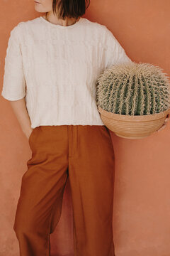 Young beautiful woman in ginger trousers and white tee holding cactus in pot against ginger wall