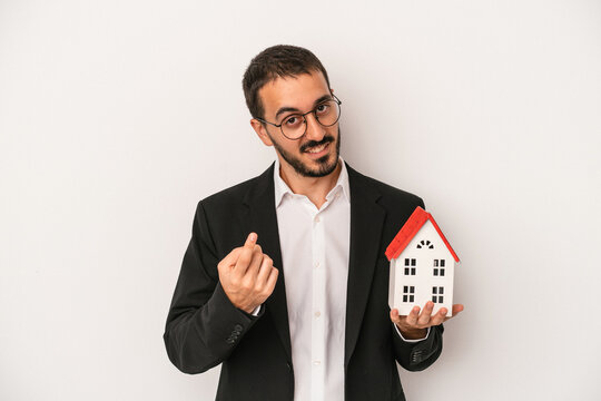Young real estate agent man holding a model house isolated on white background pointing with finger at you as if inviting come closer.