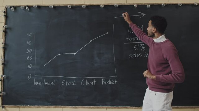 Medium of young Biracial man drawing graph with chalk on blackboard, standing indoors at daytime, talking on camera