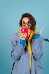 Spring autumn portrait of a woman in a blue knitted sweater, glasses, a yellow bright scarf and a gray coat, looks at the phone screen in surprise and shock