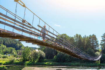 Fototapeta premium Woman with dog walks along old wooden hanging bridge on river in summer sunny day. Natural landscape. Dog on bridge
