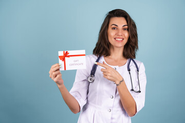 Young beautiful woman doctor in a lab coat on a blue background holds a gift certificate and smiles...
