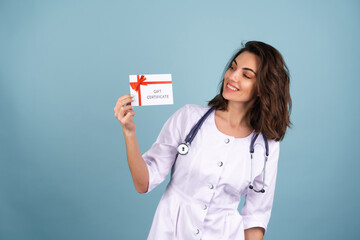 Young beautiful woman doctor in a lab coat on a blue background holds a gift certificate and smiles...
