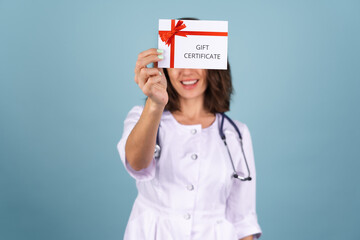 Young beautiful woman doctor in a lab coat on a blue background holds a gift certificate and smiles cheerfully