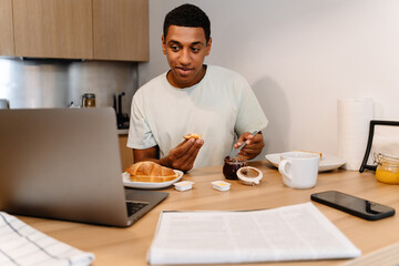 Fototapeta na wymiar Black man smiling and using laptop while having breakfast