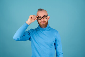 A young man with a red beard in a turtleneck and glasses on a blue background looks at the camera with a confident smile