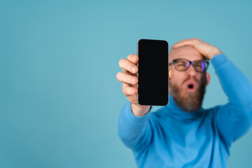 A young man with a red beard in a turtleneck on a blue background holds a phone with a blank black screen for an inscription in focus, stretches out to the camera