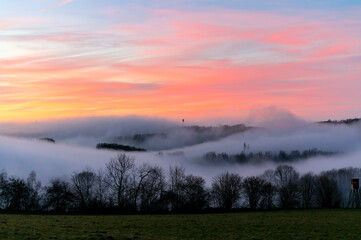Sauerland scenery panorama on a foggy winters evening after sunset. Colorful sky and wafts of mist and clouds in the valleys near Iserlohn and Altena Germany. Mystic atmosphere in rural landscape.