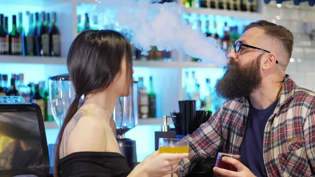A couple enjoys a date, talking and drinking alcoholic cocktails together in a stylish nightclub.A brutal bearded man smokes an electronic cigarette behind the bar.The concept of rest and love