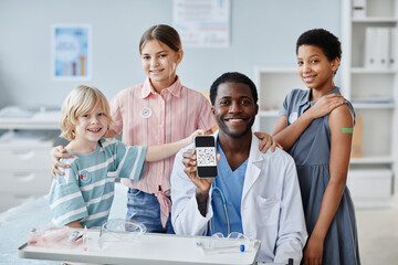 Fototapeta na wymiar Portrait of African-American doctor with group of children showing qr code in vaccination clinic and smiling at camera