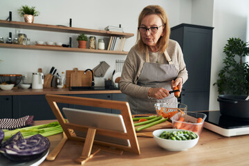 Caucasian senior woman peeling a carrot while cooking in the kitchen - obrazy, fototapety, plakaty