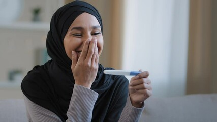 Portrait joyful surprised girl in hijab holding pregnancy test sit in living room excited with positive result beautiful islamic young woman feel happy after successful fertility treatment IVF concept