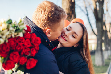 Valentines day date. Man kissing girlfriend on cheek outdoors gifting bouquet of red roses. Couple walking celebrating