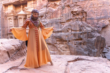 Obraz premium woman dressed in traditional clothes and headscarf staying at the top of rock on the background is Petra palace Jordan