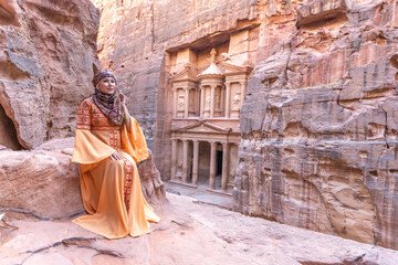 woman dressed in traditional clothes and headscarf staying at the top of rock on the background is Petra palace Jordan - obrazy, fototapety, plakaty