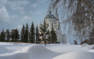 On a snowy street in the Ural village of Shurala (Ural, Russia) on a winter day. View of an old white-stone church with blue domes, surrounded by tall triangular fir trees and tall snowdrifts. 