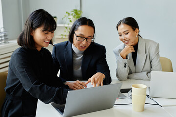 Portrait of Asian business team of three people looking at laptop screen and smiling while meeting in office