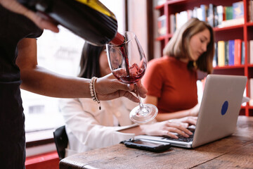 hand of a waitress pouring red wine to two freelance women sitting at the table working with the computer