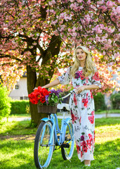spring beautiful woman in dress. girl with vintage bike. blooming sakura tree. summer fashion and beauty. lady walk in park. pink cherry tree blossom. girl carry flowers in retro bicycle