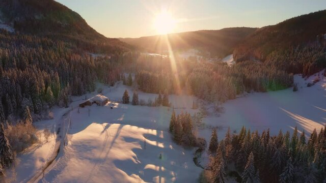 Survol image aérienne face au soleil couchant d’une vallée de montagne recouverte de neige. Lumière chaude rasante sur la foret de sapins du Jura (Valserine, Mijoux)