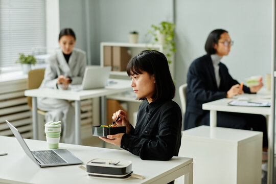 Side view portrait of young Asian woman enjoying hot lunch at desk in office, copy space