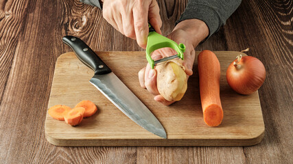 a man peels potatoes with a vegetable peeler, next to it lies a large knife, carrots and onions