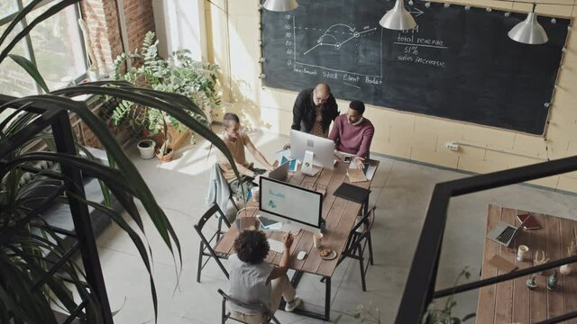 High-angle of four multiethnic colleagues using computers, sitting at table in loft-styled coworking space at daytime, working on project