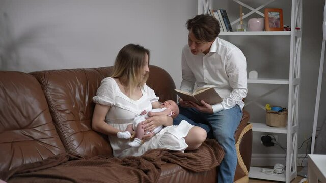 Caucasian happy father reading fairy tales to newborn son in hands of smiling woman sitting on cozy couch in living room. Man helping woman taking care of infant baby indoors. Slow motion