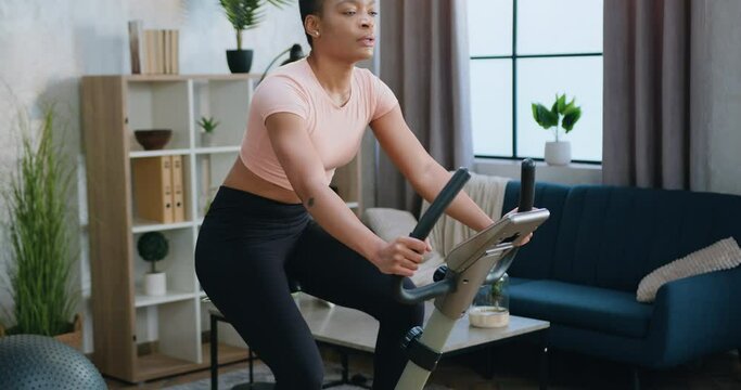 Attractive tired sporty slim young african american woman in training clothes turning the pedals on exercise bike during home workout ,active lifestyle concept