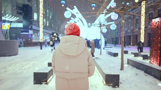 rear view camera following woman in knitted red cap and scarf walking on city square with festive colorful illuminations at night. soft snow is falling and snowflakes flying around.