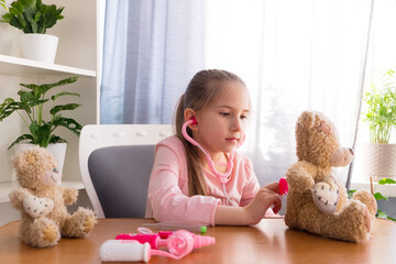 A preschool girl, dressed in a pink T-shirt, plays at home in the hospital with toys, listens to a teddy toy through a phonendoscope.