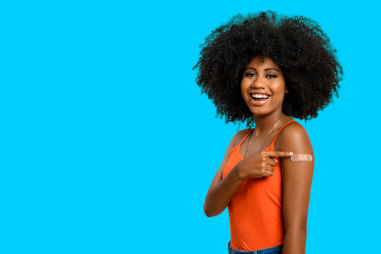 Black teen girl smile and points to her arm with vaccine sticker, she does not wear face shield, isolated on blue background.