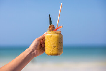 Tropical yellow mocktail drink at the beach, fresh tropical smoothy being held with the ocean in the background