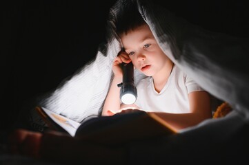 Reading book and using flashlight. Young boy in casual clothes lying down near tent at evening time.