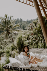 Fototapeta na wymiar A young travelling woman relaxing in the lounge area of a Bali jungle hotel surrounded by jungle, palm trees and nature