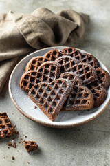 Chocolate waffles in the shape of a heart sprinkled with powdered sugar in ceramic plate on textured background with a linen napkin. Concept for valentine's day