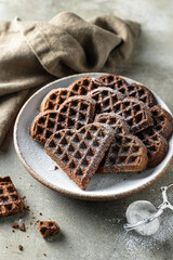 Chocolate waffles in the shape of a heart sprinkled with powdered sugar in ceramic plate on textured background with a linen napkin in the foreground, still life for valentine's day