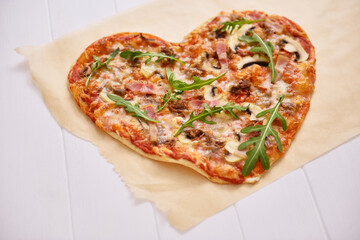 Heart-shaped pizza lies on parchment on a white wooden background, selective focus.

