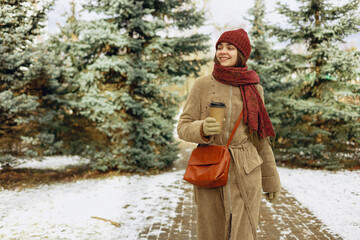 Cheerful woman walking in winter park with cup of takeaway coffee and looking away 